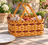 Large wood-splint picnic basket with burgundy accent rows and crossed wooden handles, styled with food on an outdoor table.